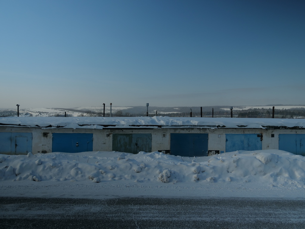 Photo of one-story storage units that have been snowed in due to plowing; a rural, snowy landscape rises above to a blue sky.