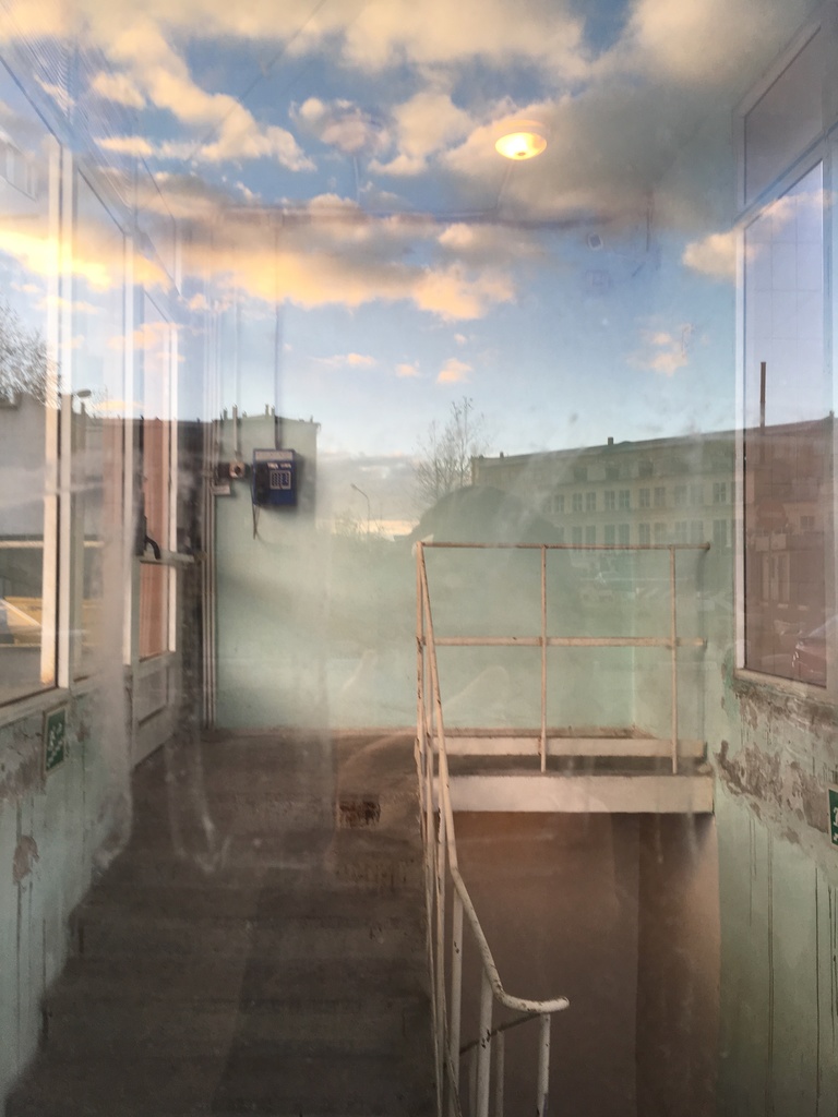 Photo of an old stairwell through a window which shows the reflection of the photographer, the blue sky, and buildings.